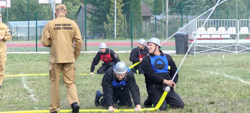 Strażacy w hełmach ćwiczą na trawie, trzymając żółty wąż strażacki. Widoczne kolorowe przeszkody i stroje. W tle płot i ławki na stadionie.