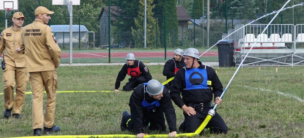 Grupa strażaków w hełmach ćwiczy na trawiastym terenie z wężami strażackimi. W tle siatka, drzewa i ławki. Dwóch strażaków w beżowych mundurach stoi z boku.