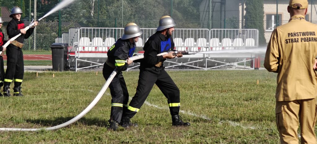 Strażacy w hełmach trzymają wąż strażacki, z którego leci woda. Mają czarne mundury z odblaskami, a w tle widoczne są trybuny i zielona trawa. Po prawej stoi osoba w jasnym uniformie z napisem PAŃST