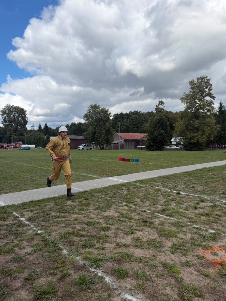 Zawodnik w żółtym kombinezonie i białym kasku biegnie po trawie. W tle są drzewa, budynki i niebo z chmurami. Na trawie widoczne są zaznaczenia toru i kolorowe wiadra.