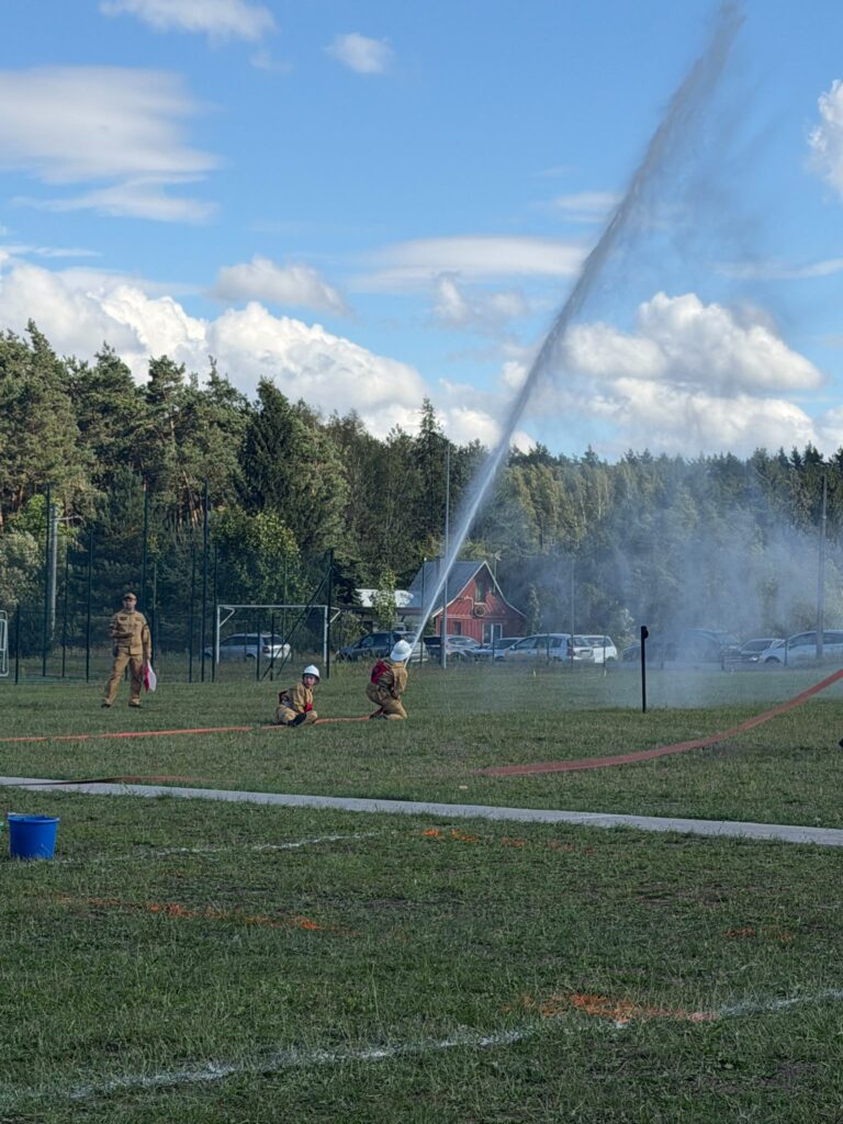 Na zdjęciu strażacy w hełmach rozkładają wąż strażacki. Tło stanowi niebieskie niebo i zielony las. Woda z węża tworzy wysoką fontannę.