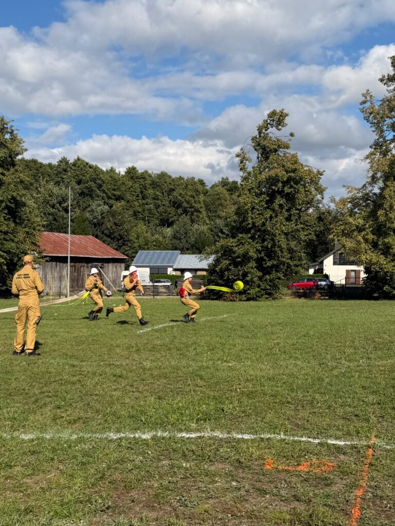 Grupa osób w żółtych uniformach biegnie po trawie z wężem strażackim. W tle drzewa, budynki oraz niebieskie niebo z chmurami. Na ziemi widać białe i pomarańczowe linie.