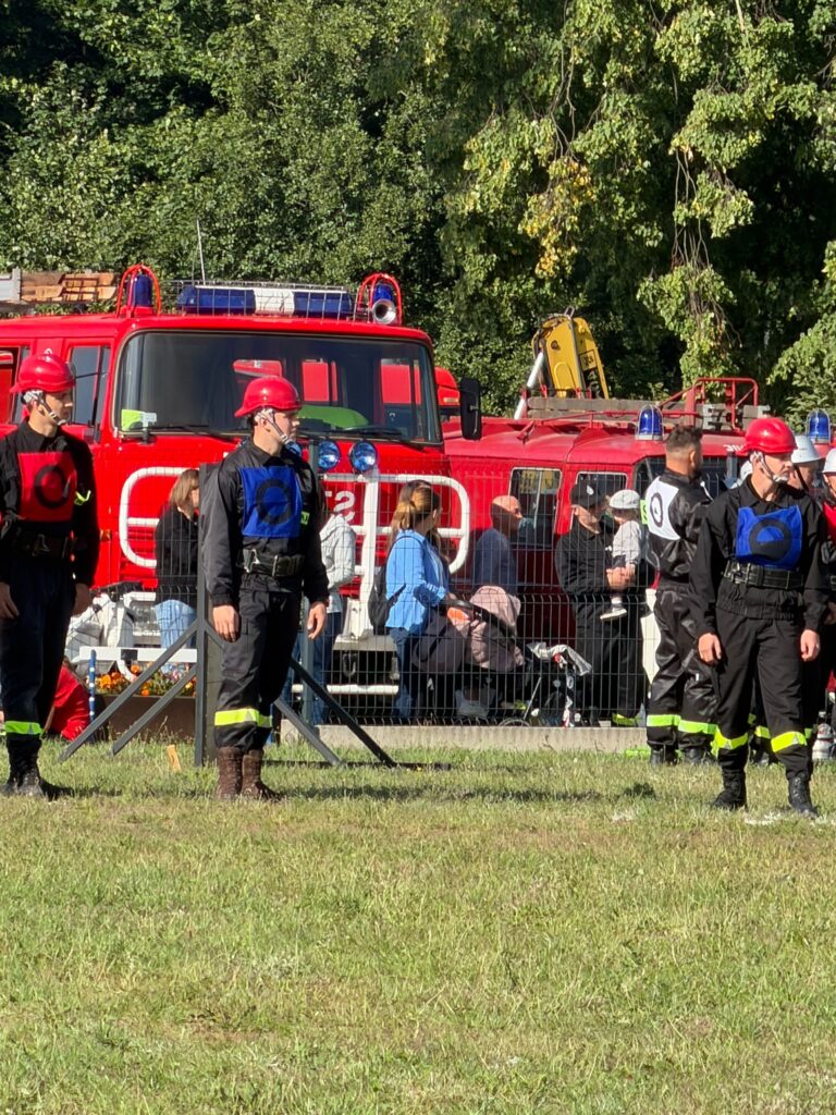 Strażacy w czarnych mundurach i czerwonych kaskach stoją na trawie przed czerwonymi wozami strażackimi. Tło stanowią zielone drzewa. Obok widać ludzi za ogrodzeniem.
