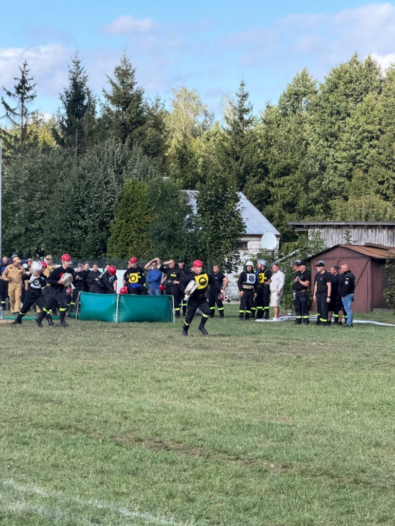 Grupa strażaków w czerwonych kaskach biegnie po trawie. W tle widoczni są obserwatorzy i drzewa. Słoneczny dzień, jasne niebo.