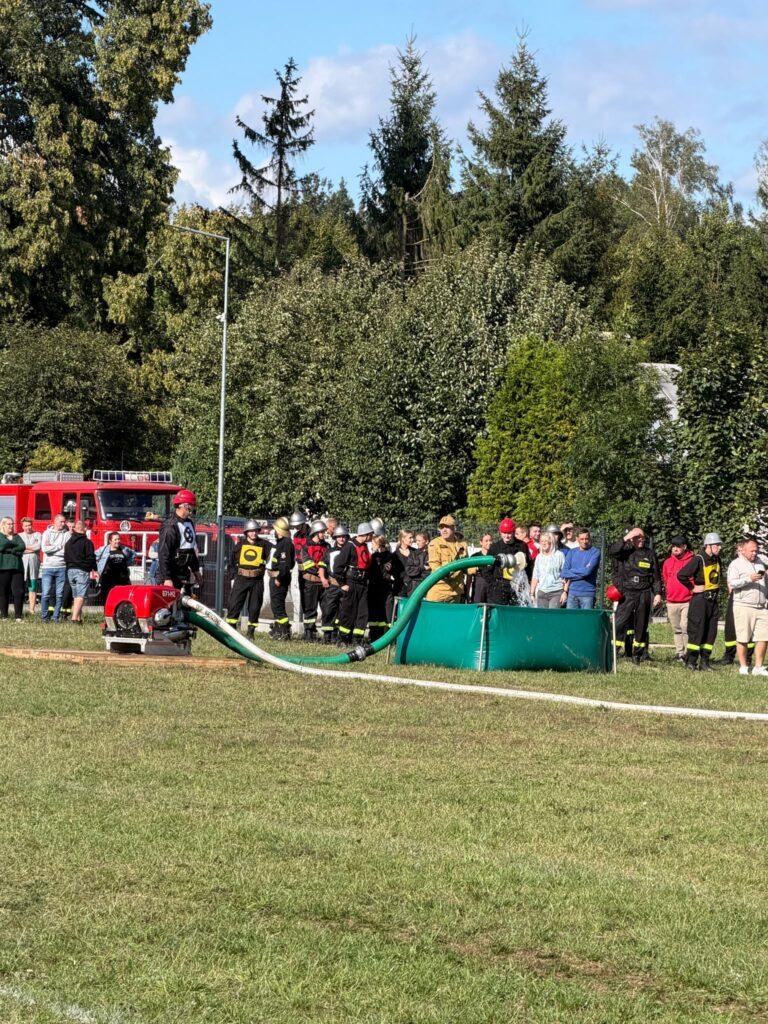 Na zdjęciu strażacy w czarnych mundurach z hełmami przy sprzęcie pożarniczym. W tle widoczna jest czerwona strażacka ciężarówka i zielone drzewa. Obok stoi grupa obserwujących ludzi.