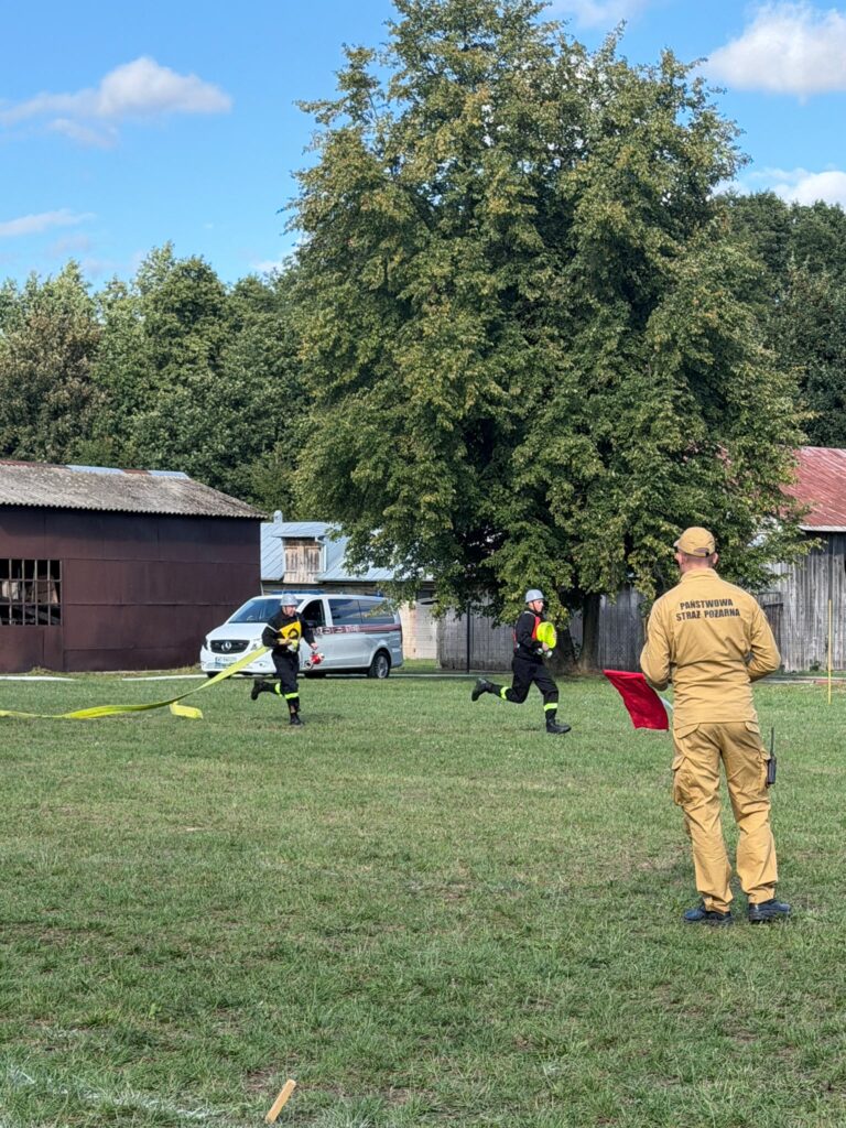Strażacy w czarnych mundurach biegną z żółtym wężem strażackim po trawie. Na pierwszym planie stoi osoba w żółtym stroju z czerwoną flagą. W tle zielone drzewa i brązowy budynek pod niebieskim niebem.