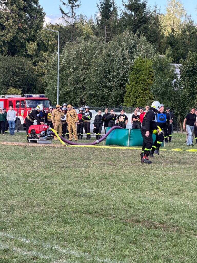 Grupa strażaków w strojach ochronnych pracuje przy sprzęcie na trawiastym terenie. W tle stoi czerwony wóz strażacki, a za nim widoczny jest tłum obserwujący wydarzenie. Otoczenie to zielone drzewa, a niebo jest pogodne.