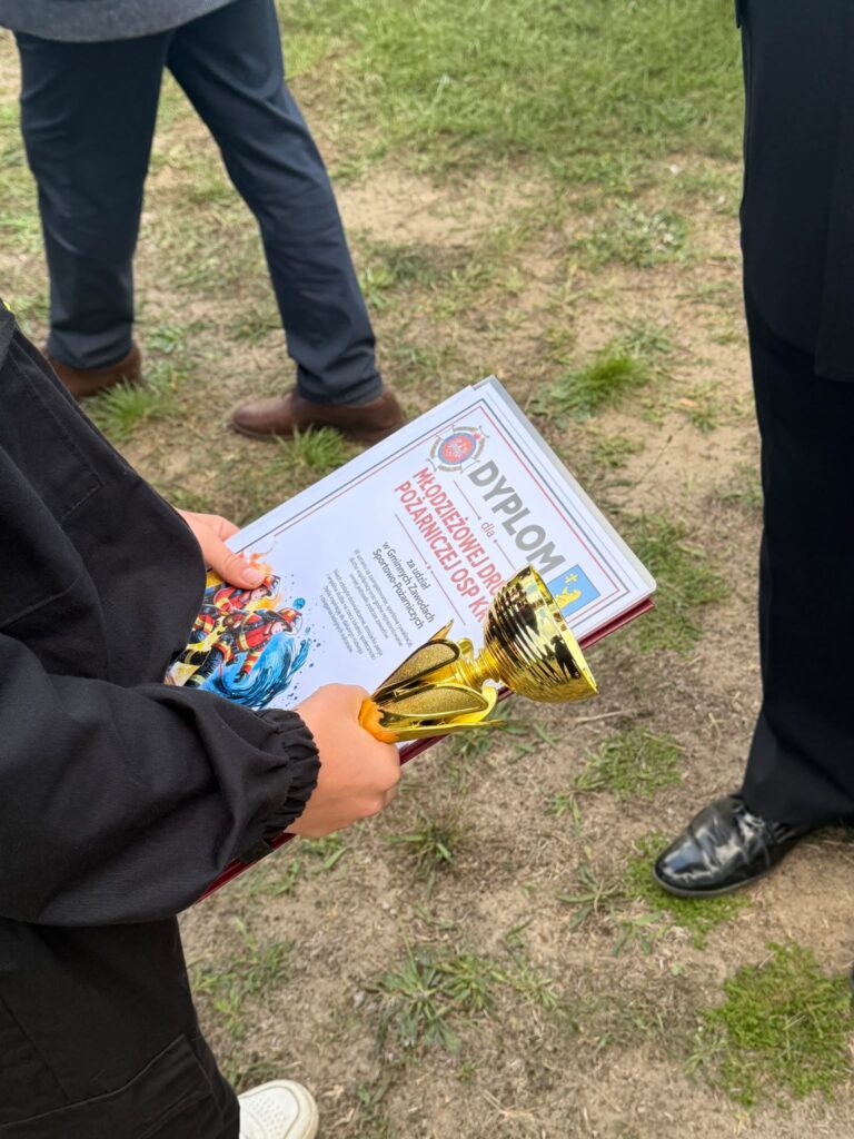 Na zdjęciu osoba trzyma złoty puchar i dyplom. Tło jest trawiaste, z postaciami w ciemnym ubraniu. Dyplom ma czerwony napis i kolorowe grafiki.