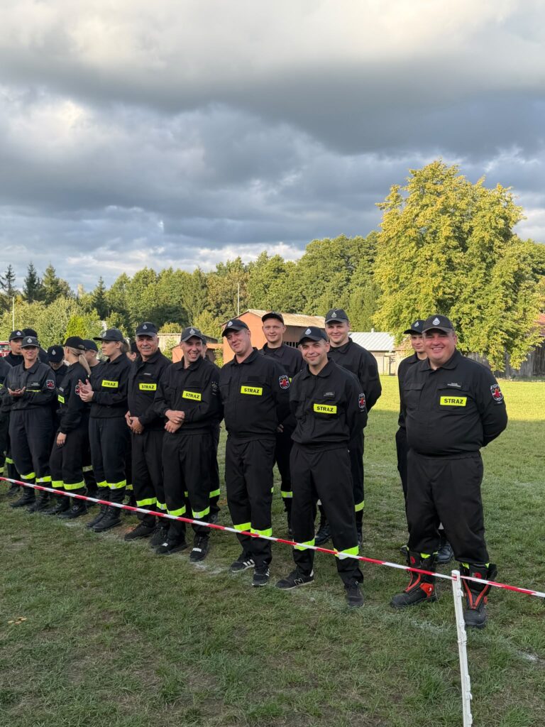 Grupa strażaków w czarnych mundurach stoi na trawie, uśmiechając się. Mają żółte napisy STRAŻ na klatkach piersiowych. W tle drzewa i chmury.