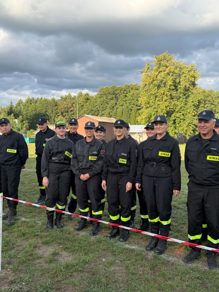 Grupa strażaków w czarnych mundurach stoi na trawie w parku. W tle widać zielone drzewa i budynki. Niebo pokryte jest ciemnymi chmurami.