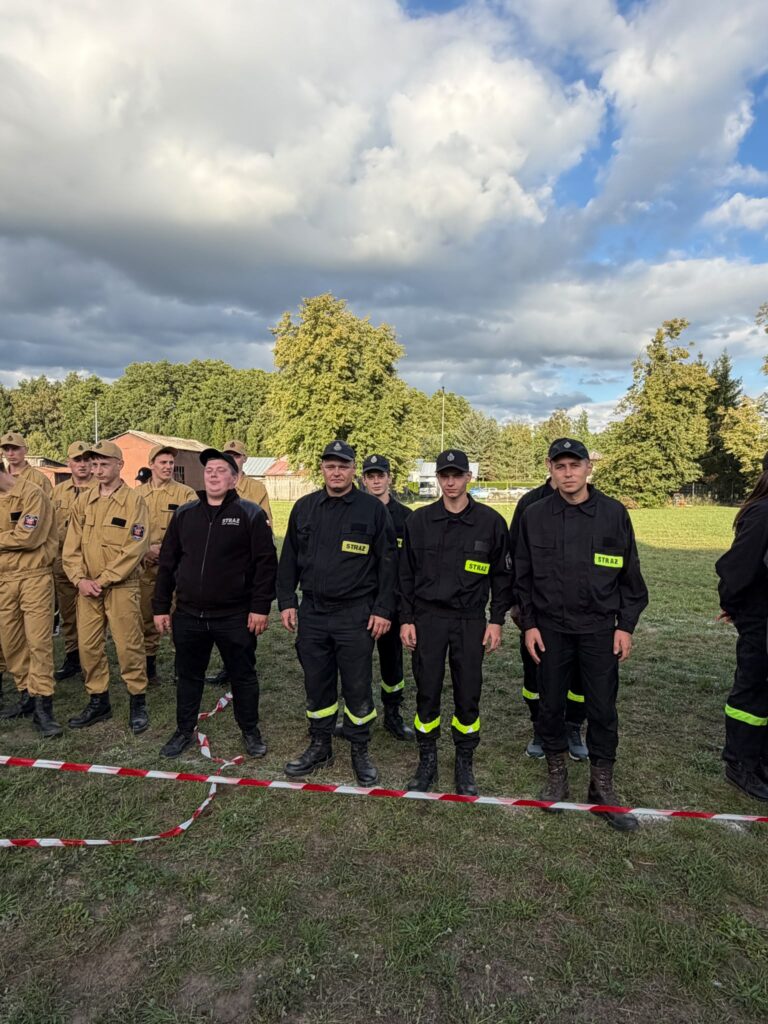 Grupa ludzi w mundurach strażackich stoi na trawniku. Tło to drzewa i pochmurne niebo. Osoby są w czarnych i beżowych strojach.