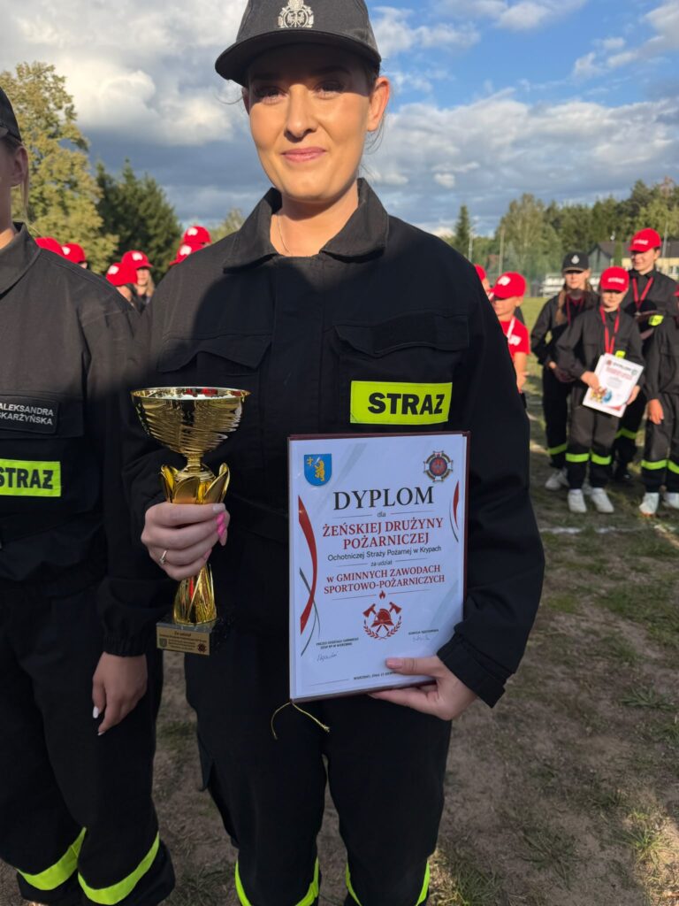 Na zdjęciu kobieta w czarnym mundurze strażackim trzyma puchar i dyplom. W tle grupa strażaków w czerwonych czapkach. Niebo jest pochmurne, a otoczenie zielone.