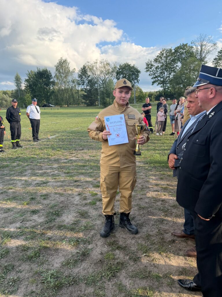 Na zdjęciu młody mężczyzna w beżowym mundurze trzyma dyplom i puchar. W tle trawiasta polana i kilka osób. Niebo jest niebieskie z chmurami.