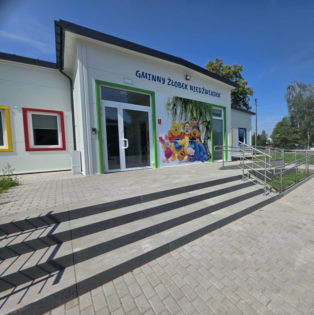 Budynek żłobka z jasnymi ścianami, kolorowe okna. Na frontowej ścianie wesoły mural z postaciami. Przed wejściem schody i podjazd.