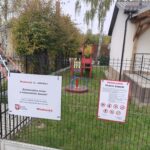 74788 Obraz przedstawia świeżo wybudowany plac zabaw w miejscowości Jaworek. Widać czerwone i niebieskie elementy zabawowe, a w tle zieleń drzew i budynek. Dwa informacyjne banery zawierają zasady korzystania z placu oraz informacje o projekcie.