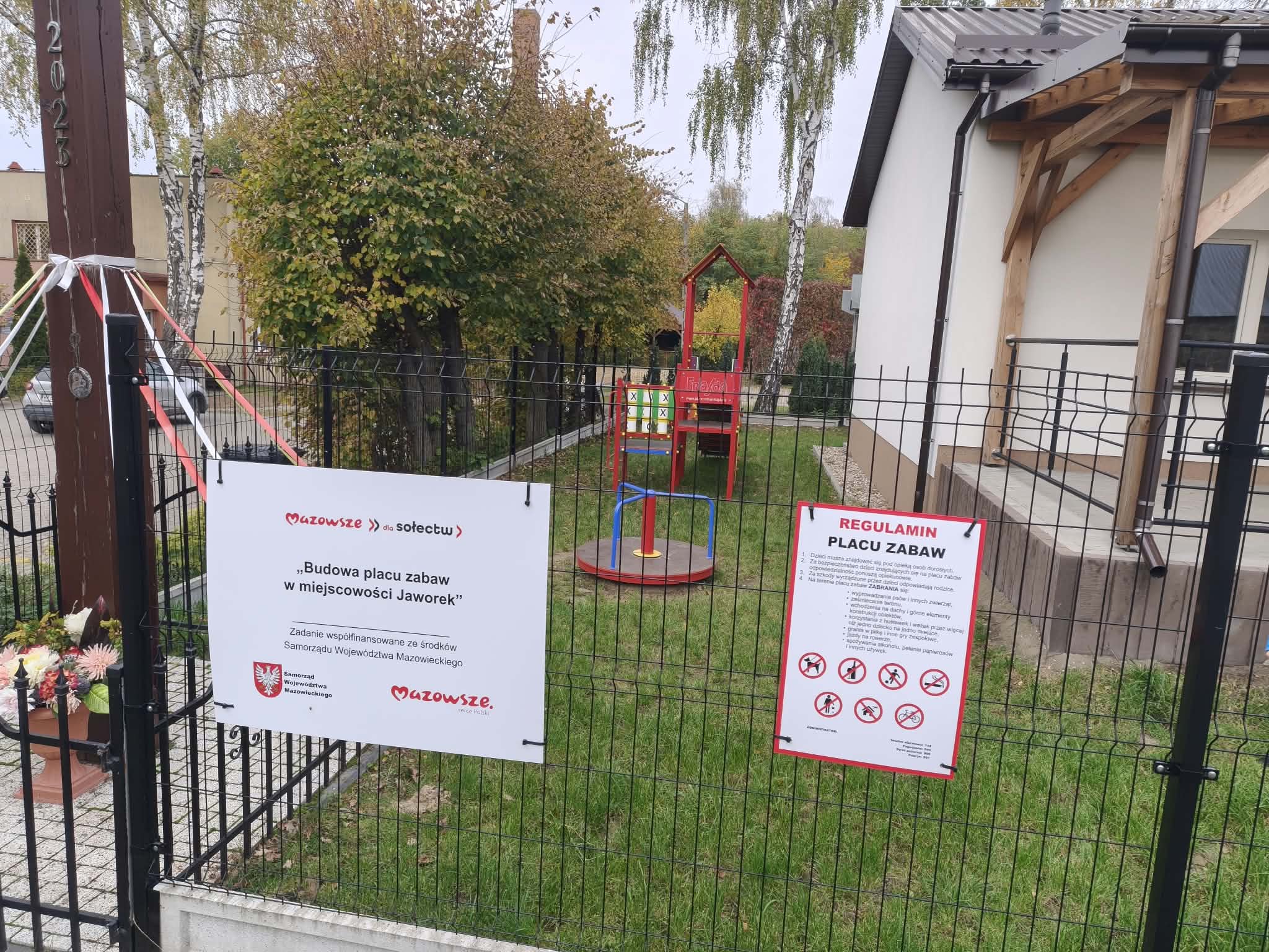 Obraz przedstawia świeżo wybudowany plac zabaw w miejscowości Jaworek. Widać czerwone i niebieskie elementy zabawowe, a w tle zieleń drzew i budynek. Dwa informacyjne banery zawierają zasady korzystania z placu oraz informacje o projekcie.
