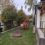 74790 Na zdjęciu widoczny jest kolorowy plac zabaw w ogrodzie, zdominowany przez zieleń trawy i drzew. W centralnej części znajduje się czerwona huśtawka oraz zestaw zabawowy w kolorach czerwonym, niebieskim i żółtym. Tło obrazka tworzą dom oraz wyżłobiona przestrzeń, co podkreśla przytulną atmosferę miejsca.