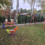 74791 Na zdjęciu widać kolorowy plac zabaw z trzema atrakcjami: huśtawką, konikiem na sprężynie oraz ławką. Otoczenie jest zielone z broszkami drzew i krzewów, a w tle widoczna jest siatka oraz budynki. Całość utrzymana w stonowanych kolorach, z domieszką intensywnych barw na zabawkach.