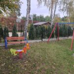 74792 Obraz przedstawia kolorowy plac zabaw z huśtawką i zjeżdżalnią w otoczeniu zieleni. Na pierwszym planie widoczna jest wesoła huśtawka w czerwono-niebieskich kolorach oraz sprężynowy koń w zielono-żółtej tonacji. W tle rosną drzewa, a cały teren jest otoczony płotem, co nadaje przytulny charakter.