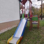 74793 Na zdjęciu znajduje się kolorowy plac zabaw z metalową zjeżdżalnią. Struktura jest pomalowana na czerwono, zielono i niebiesko, a zjeżdżalnia ma błyszczący, srebrny kolor. Tło stanowi trawa oraz biały budynek, a wszystko otoczone jest drzewami w jesiennych barwach.