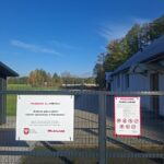 Na zdjęciu widać ogrodzenie terenu budowy placu zabaw i siłowni w Karczewcu. Tło stanowią niebieskie niebo oraz zielone drzewa. Widoczne są także dwa informacje: o budowie oraz regulamin placu zabaw, umieszczone na białych tablicach.