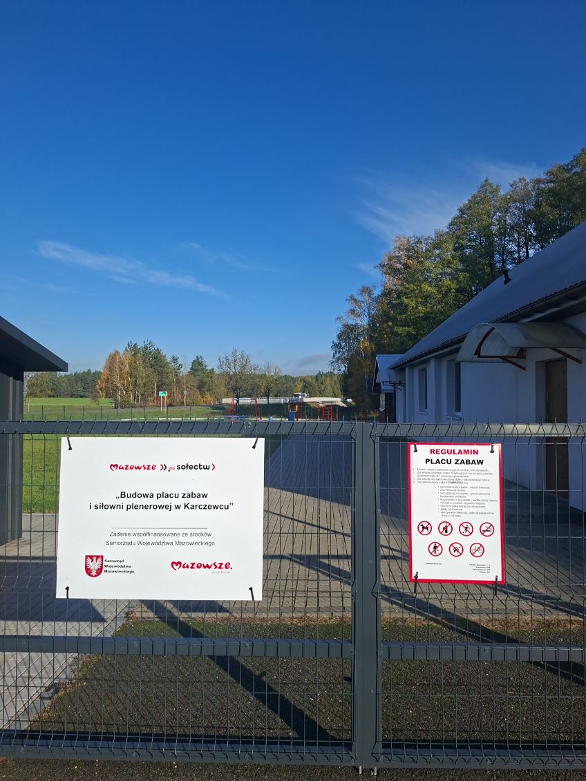 Na zdjęciu widać ogrodzenie terenu budowy placu zabaw i siłowni w Karczewcu. Tło stanowią niebieskie niebo oraz zielone drzewa. Widoczne są także dwa informacje: o budowie oraz regulamin placu zabaw, umieszczone na białych tablicach.